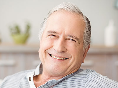 The image shows a smiling older man with gray hair, wearing a dark shirt and glasses, sitting in a relaxed posture with his hands on his legs. He appears content and at ease.